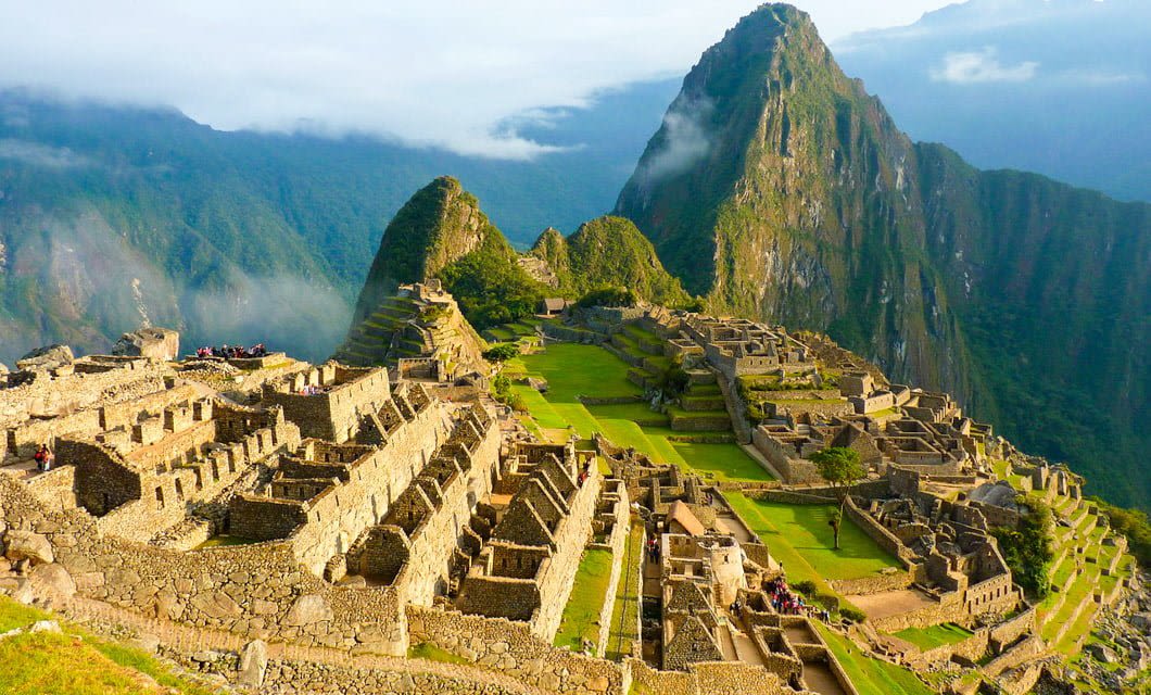 Ancient stone ruins of Machu Picchu sit on a lush green mountain ridge, inviting adventurers on a Coast to Coast Tour through steep, forested peaks and misty clouds under a bright sky.