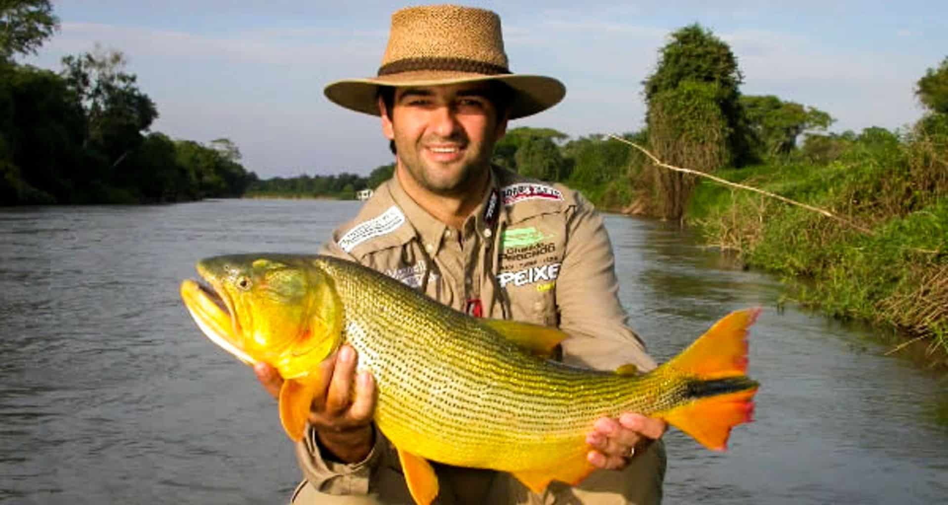 A smiling man wearing a wide-brimmed hat and fishing gear holds a large, yellow-striped fish beside a river in Brazil’s lush Pantanal, surrounded by green trees and bushes on an unforgettable Fishing Tour.
