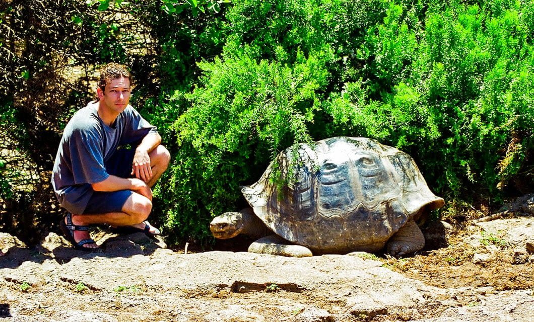 A man squats next to a very large Galapagos tortoise on a dirt path, with lush green bushes in the background under bright sunlight—perfect for an unforgettable travel moment on the Galapagos Loop.