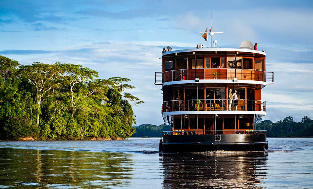 A modern, three-deck river cruise ship travels along a wide, calm river bordered by lush green Ecuadorian Rainforest under a blue sky with clouds—an unforgettable Amazon Explorer experience.