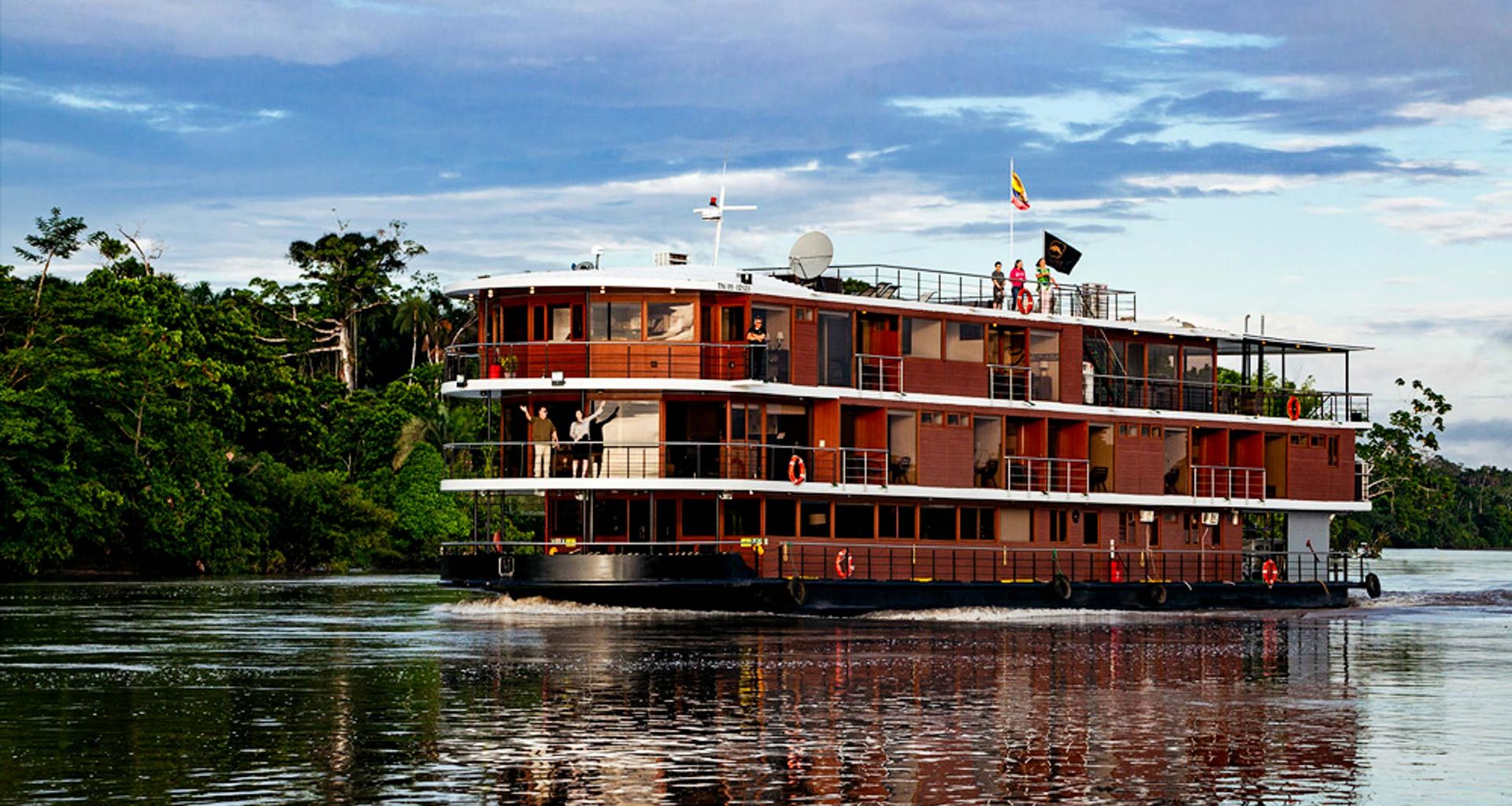 The Manatee Amazon Explorer, a large modern riverboat, cruises along a wide river in the Ecuadorian Rainforest. Surrounded by lush jungle and a partly cloudy sky, several people stand on the top deck as flags fly from the boat.