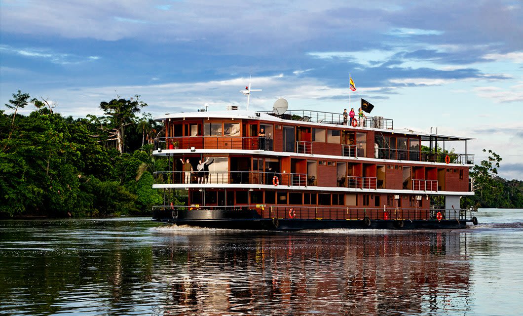A modern, multi-level riverboat with wooden paneling cruises on a calm, reflective river in the Ecuadorian Rainforest. People stand on the top deck as several flags fly above during their Amazon cruise beneath a blue sky and lush jungle canopy.