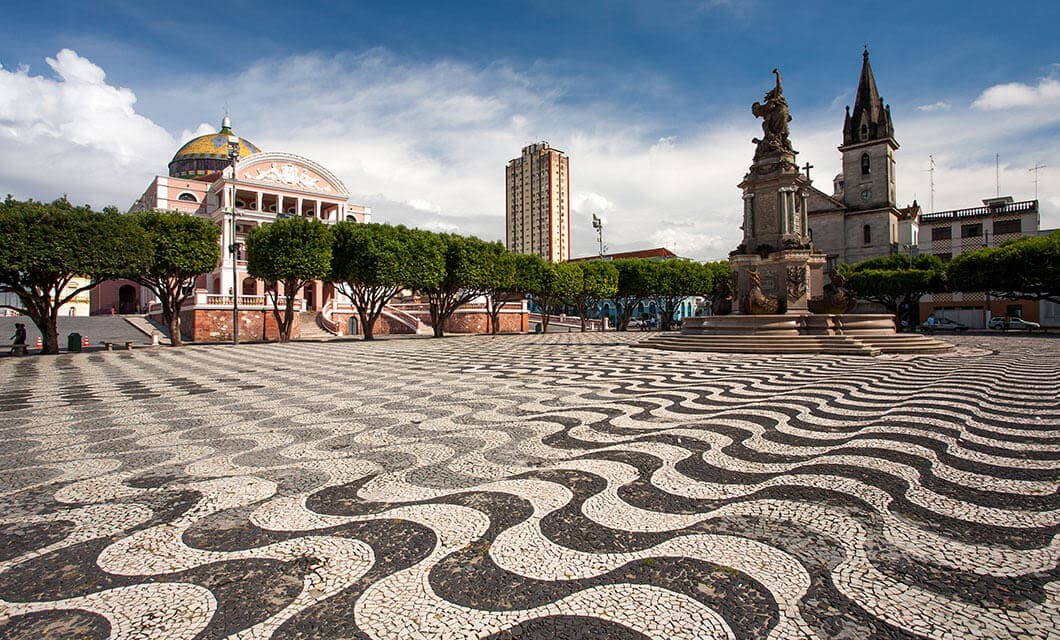 A large square with a wavy black-and-white patterned stone pavement, a statue on a fountain, and trees lining the area. Colorful buildings and a domed theater evoke scenes from a Remote Amazon Tour under a blue sky with clouds.