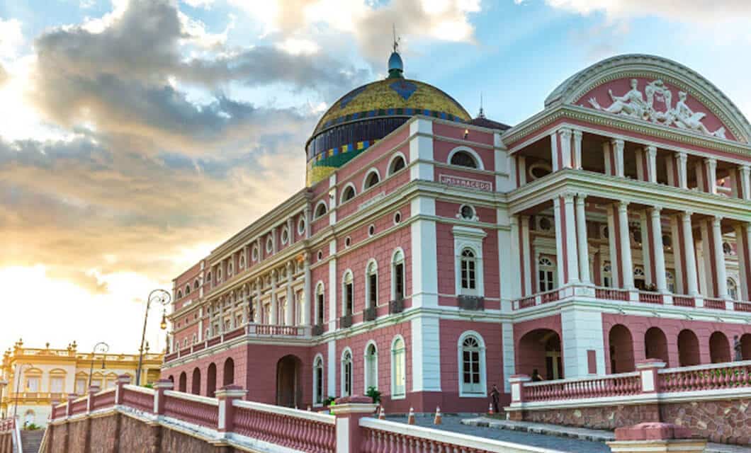 A historic pink and white neoclassical building with columns and a colorful dome, viewed during sunset on a Remote Amazon Tour; the sky is partly cloudy, and the building is surrounded by a balustrade.