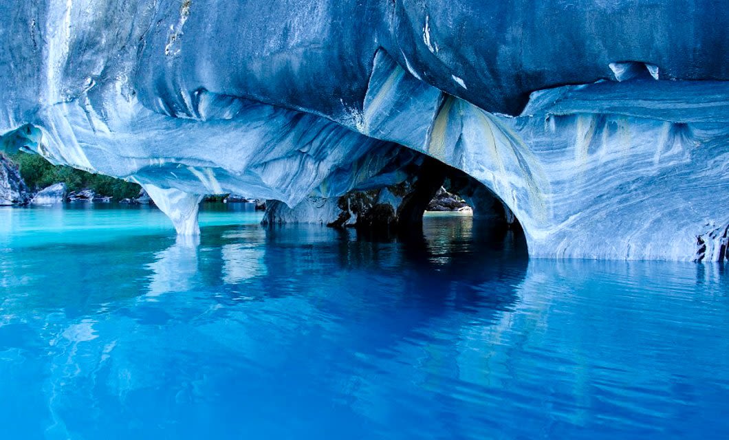 Marble caves with smooth, swirling blue and white patterns rise above vibrant turquoise water on the Patagonian Route 40, reflecting the rock formations. Some greenery is visible in the background.