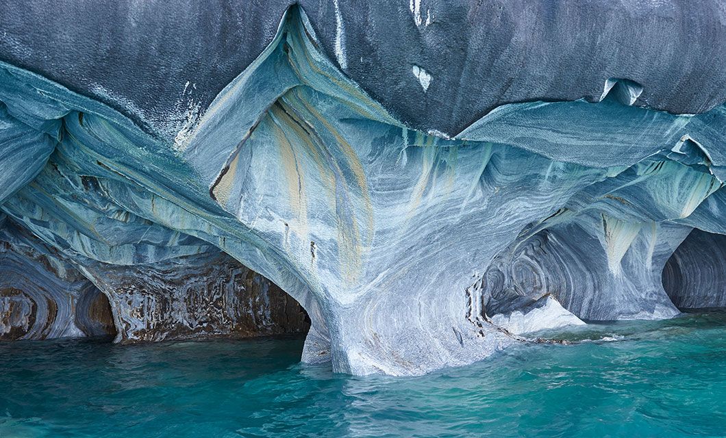 Smooth, swirling marble rock formations with blue, gray, and beige patterns rise above clear turquoise water in a cave-like setting along the Carretera Austral—an iconic stop on any Patagonia tour.
