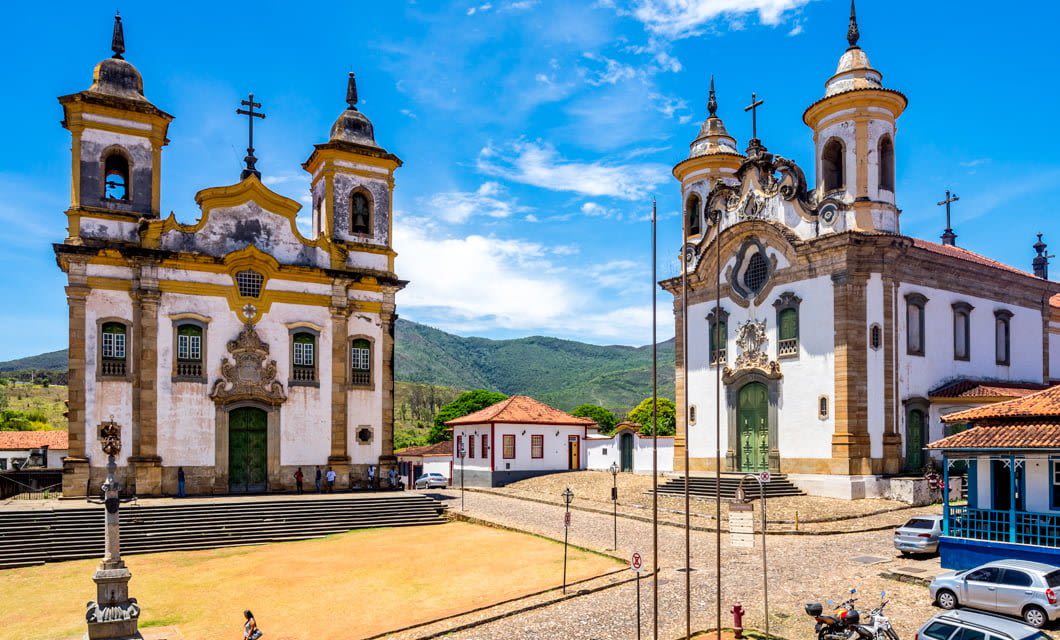 Two ornate Baroque churches with twin bell towers face a historic town square, part of the Brazil Gold Route, with mountains and blue sky in the background. Some cars and people are visible near the buildings.