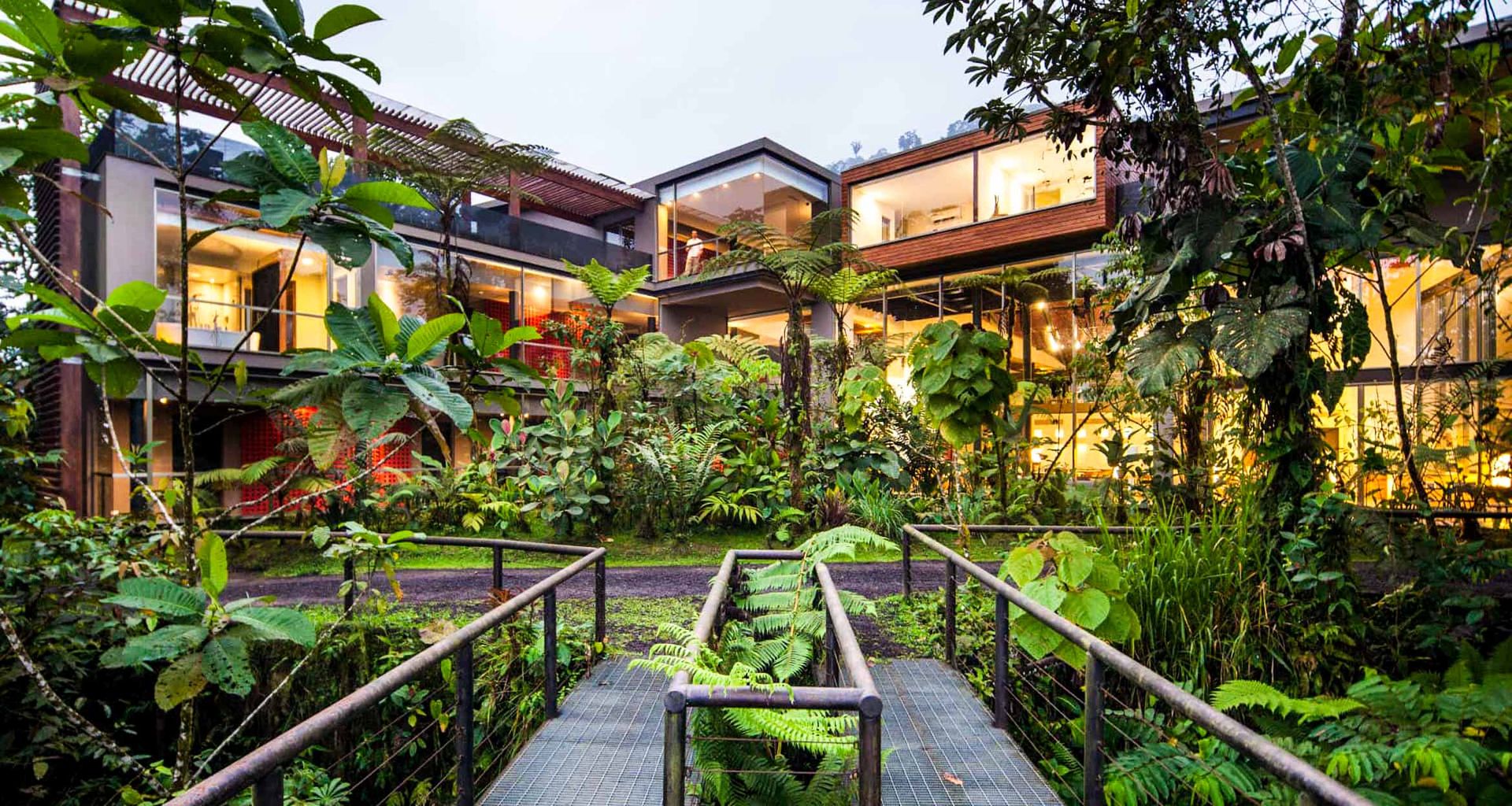 A modern glass and wood building surrounded by lush tropical plants and trees in the Ecuador rainforest, viewed from a metal walkway leading through dense greenery toward the brightly-lit Mashpi Lodge.