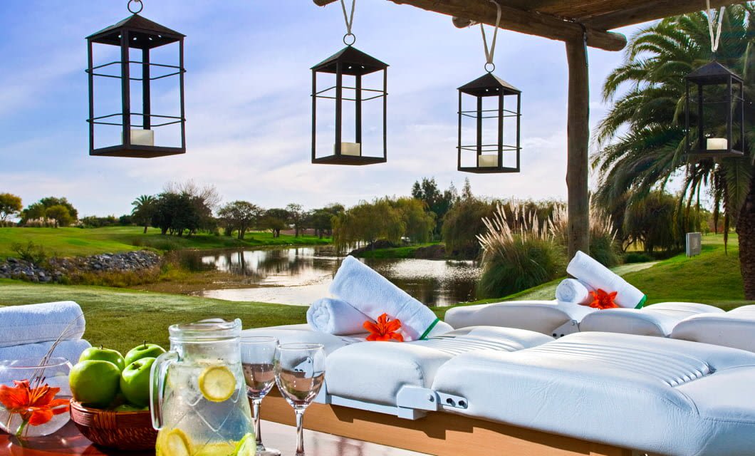 A relaxing outdoor spa setup with white lounge chairs, rolled towels, and lanterns hanging from a pergola overlooks a scenic pond and palm trees—an ideal retreat after an Argentina golf or South America golf tour.