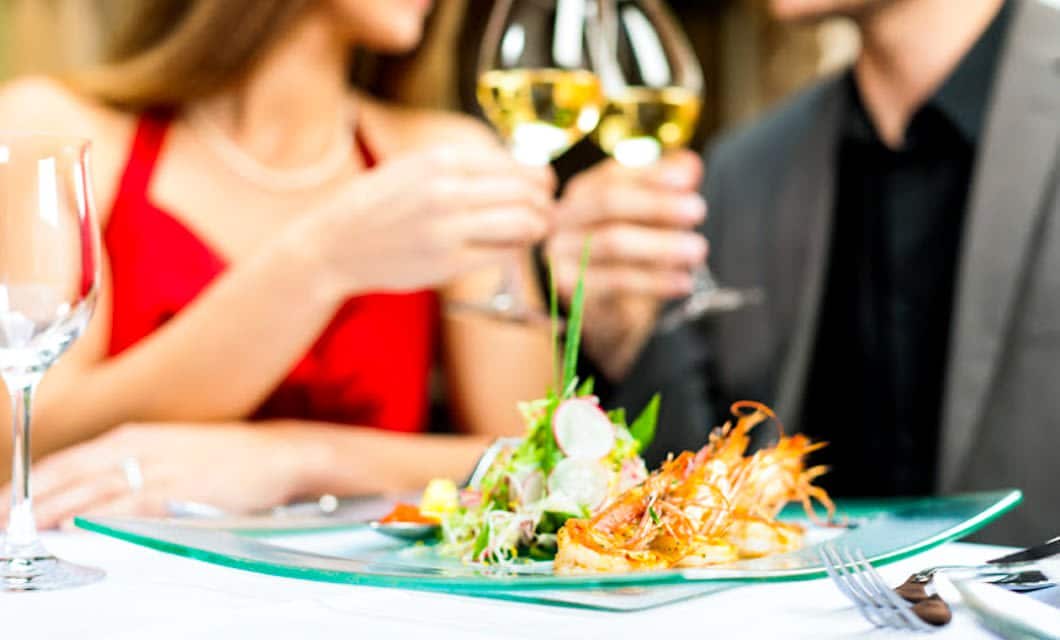 A couple dressed elegantly clinks glasses of Mendoza wine at a restaurant table, with a gourmet seafood dish and salad in focus in the foreground. The mood appears romantic and celebratory.