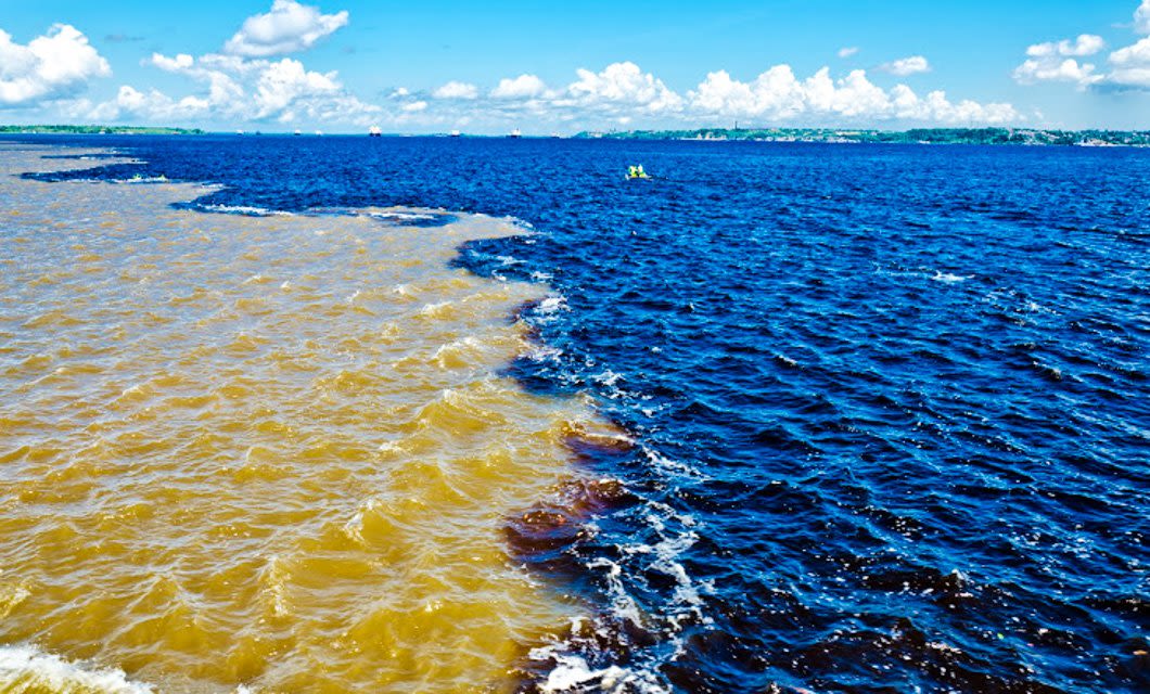 Two rivers with distinctly different colors, one muddy brown and the other dark blue, flow side by side without immediately mixing under a bright blue sky—an iconic sight on a Deluxe South America Tour.