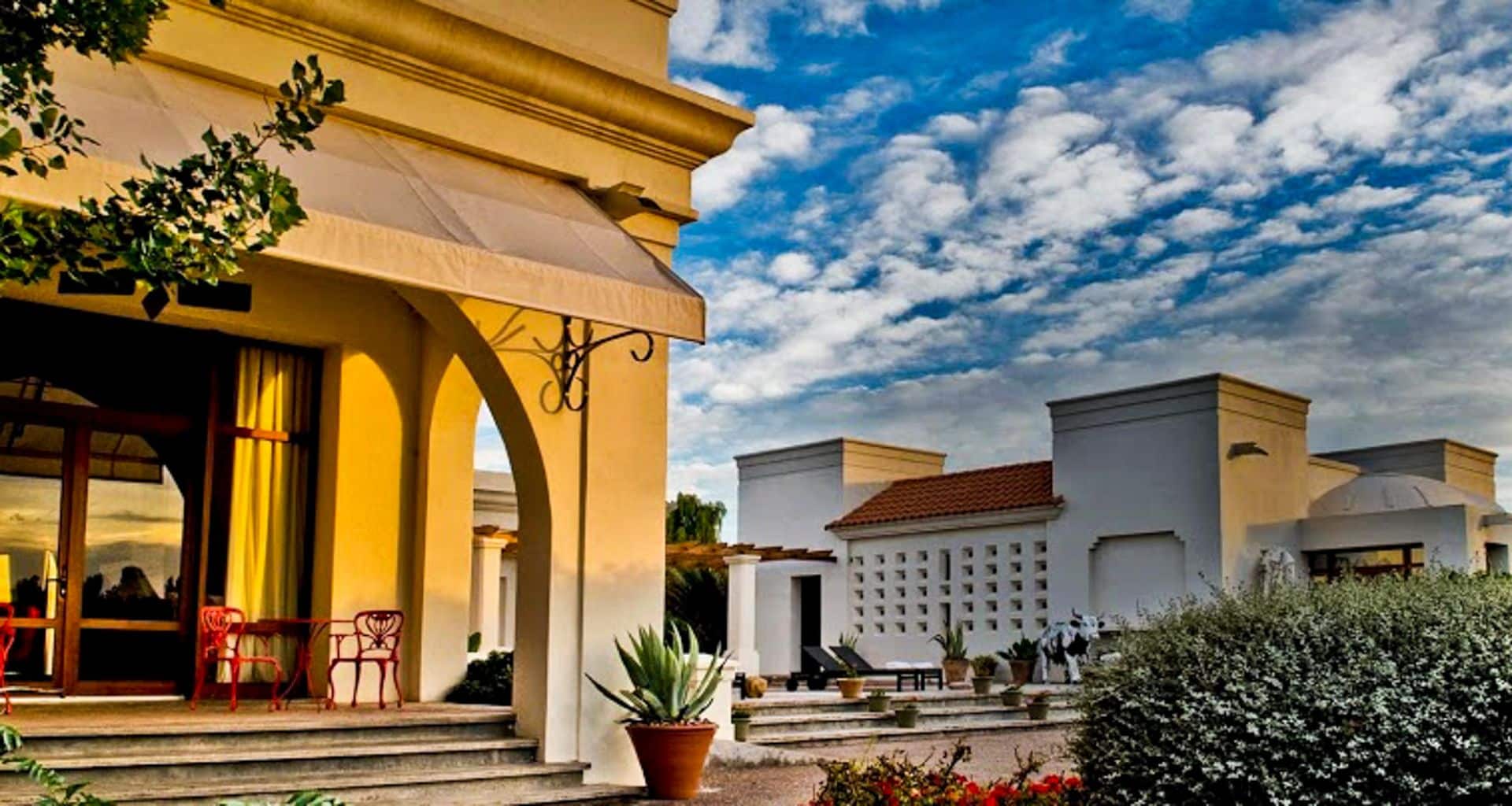 A sunlit villa with arched doorways, red chairs, and potted plants stands in the foreground at Cavas Wine Lodge, while modern white buildings and lush greenery are visible under a bright, partly cloudy sky.
