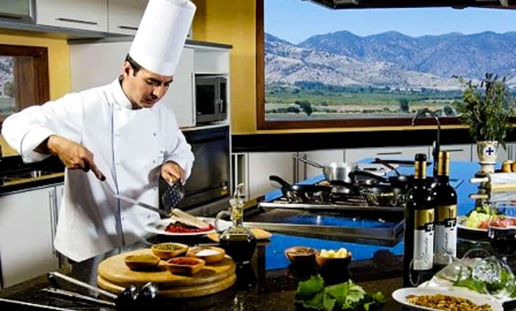 A chef in a white uniform and hat prepares food in a modern Mendoza kitchen with large windows revealing a scenic mountain view—a perfect setting for a romantic honeymoon, complete with ingredients, wine bottles, and cooking utensils on the counter.