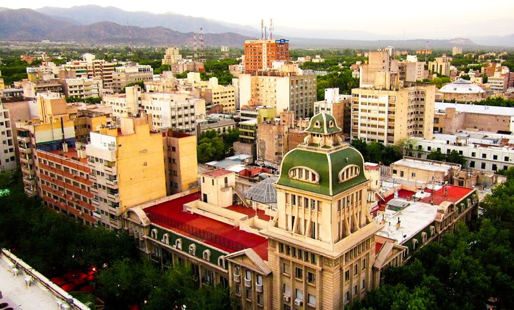 A cityscape in South America with mid-rise buildings, green trees, and a prominent historic building with a green dome roof in the foreground. Mountains are visible in the hazy background—perfect for starting a memorable wine tour.