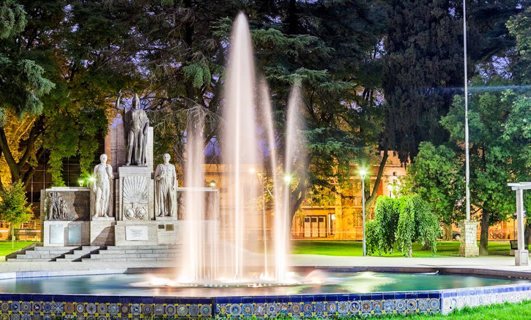 A lit fountain at night in front of statues and trees in a park, with soft lights illuminating the surrounding area—reflecting the beauty and diverse landscapes of Argentina, the Land of Contrast.