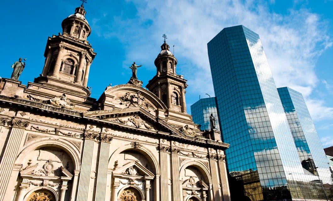 Historic stone cathedral with ornate towers stands in contrast to modern glass skyscrapers under a bright blue sky, much like the striking architectural contrasts found on a Patagonia Overland adventure through Northern Patagonia.