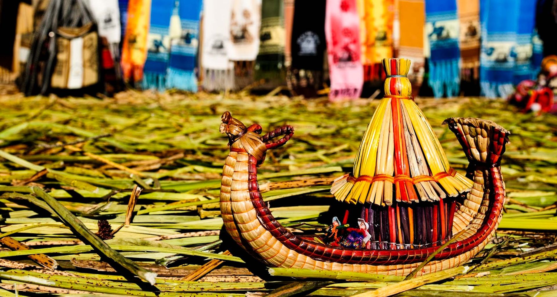 A colorful reed boat model with a thatched roof sits on a bed of dried reeds, evoking the spirit of Travels of the Incas, as vibrant woven textiles hang in the blurred background.