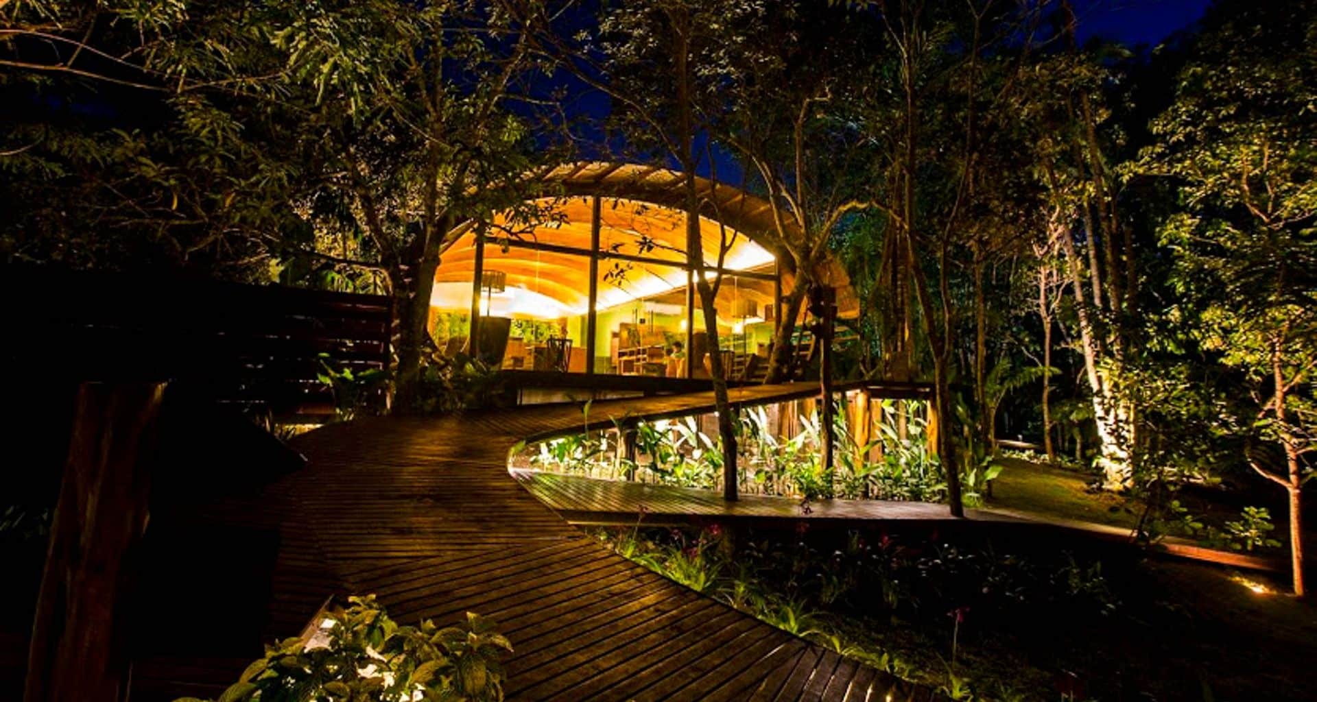 A modern, wooden treehouse with large glass windows glows warmly at night, nestled in the lush Amazon rainforest at Mirante do Gavião and connected by a curved wooden walkway to this unique Amazon Lodge.
