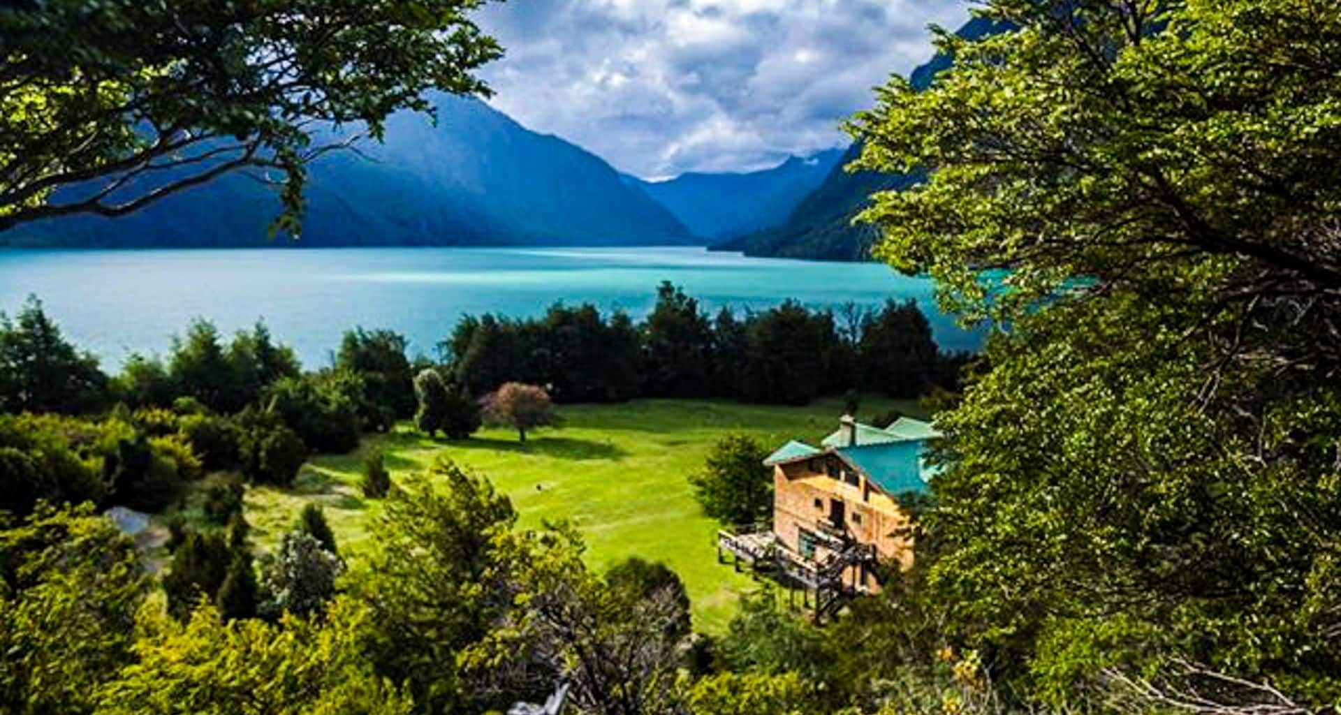 Mitico Puelo Lodge is a wooden house surrounded by trees near a grassy lawn, with a large turquoise lake and forested mountains in the background under a partly cloudy sky.