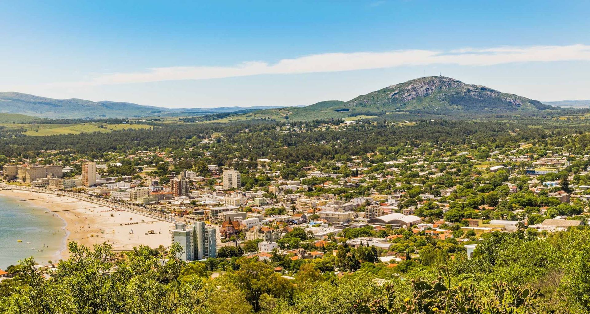 A coastal city like Piriapolis, with sandy beaches, high-rise buildings, and dense greenery, nestles below rolling hills and a large mountain under a clear blue sky.