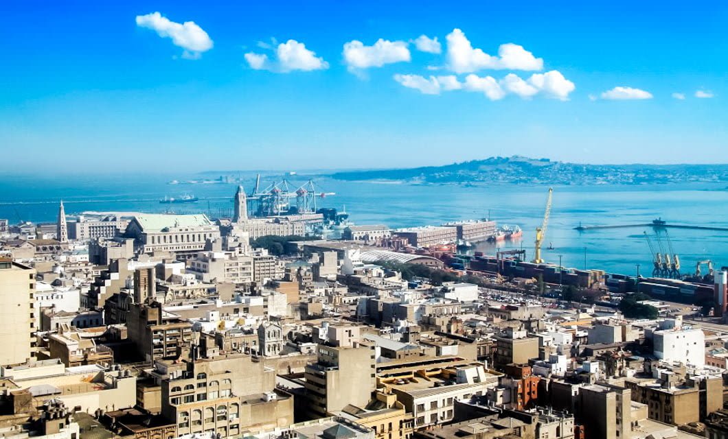 Aerial view of Buenos Aires, a coastal city where the urban rhythm meets a busy port with cranes and ships, set against a calm blue sea under a bright sky. Distant hills frame this vibrant scene.