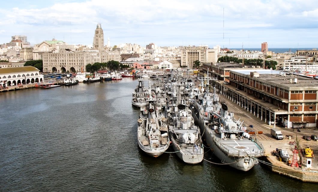 A busy harbor in Montevideo with multiple naval ships docked at piers, surrounded by historic and modern buildings under a partly cloudy sky. The cityscape of Uruguay stretches into the background.