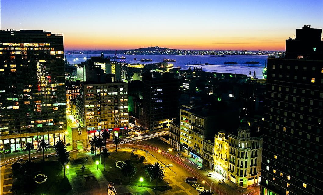 A vibrant Montevideo cityscape at dusk shows illuminated buildings, busy streets, and palm trees in the foreground, with a calm bay and ships visible under a colorful sunset sky in the background.