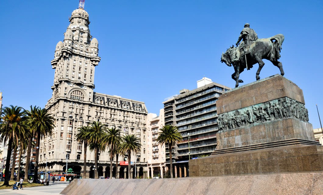 A large equestrian statue on a stone pedestal stands in front of the historic Palacio Salvo and palm trees in Plaza Independencia, Montevideo, Uruguay, under a clear blue sky—a must-see before visiting Punta del Este or Colonia.