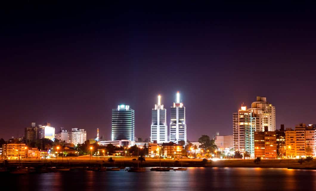 Nighttime cityscape of Montevideo with illuminated high-rise buildings and their lights reflecting on the water. The sky is dark, and the city glows with warm and cool tones—perfect for experiencing an unforgettable Uruguay Tour.