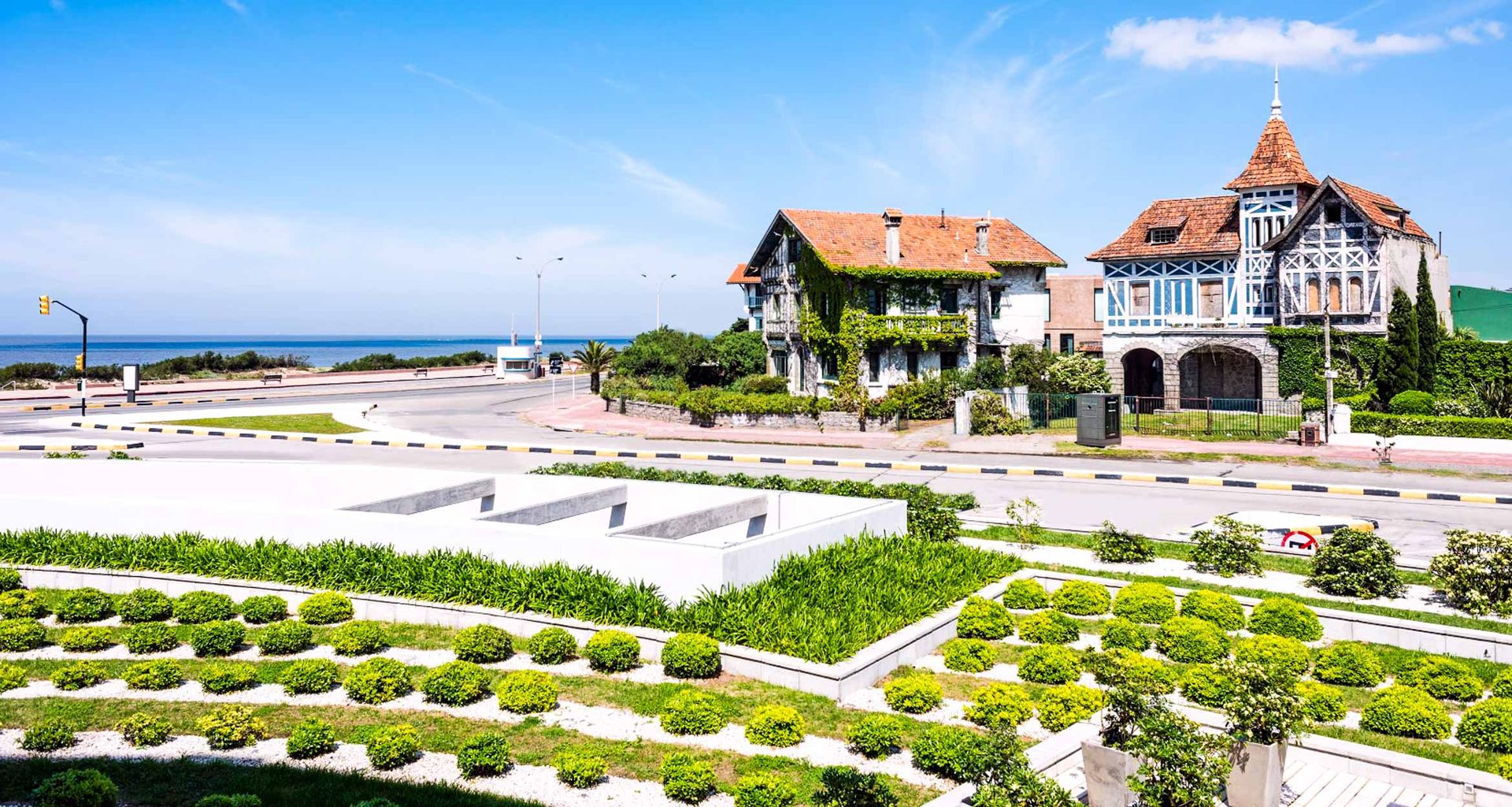 A scenic view of a coastal road with two charming, historic houses featuring red-tiled roofs and decorative facades—perfect for an Uruguay tour. Manicured gardens sit in the foreground, with the sea visible beyond under a clear blue sky.