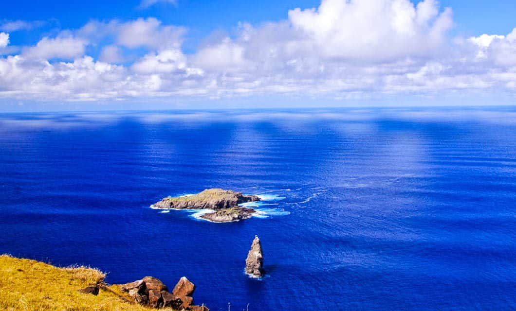 A vibrant blue ocean stretches to the horizon under a partly cloudy sky, with two small rocky islands and a sea stack reminiscent of Easter Island rising from white waves. Yellow grass and rocks appear in the foreground.