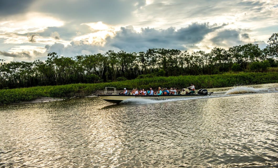A motorboat with several passengers speeds along a river, creating a trail of water. Dense green trees line the riverbank under a cloudy sky, reminiscent of an adventurous Amazon cruise with sunlight breaking through.