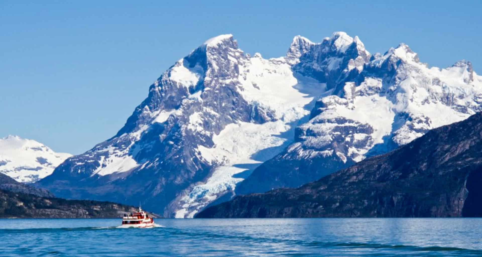 A small red and white boat sails on a blue lake in Argentine Patagonia, with towering, snow-covered mountains and a glacier in the background under a clear blue sky.