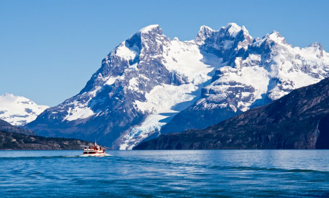 A small red and white boat cruises on a blue lake in Argentine Patagonia, with massive snow-capped mountains and a glacier in the background under a clear blue sky.