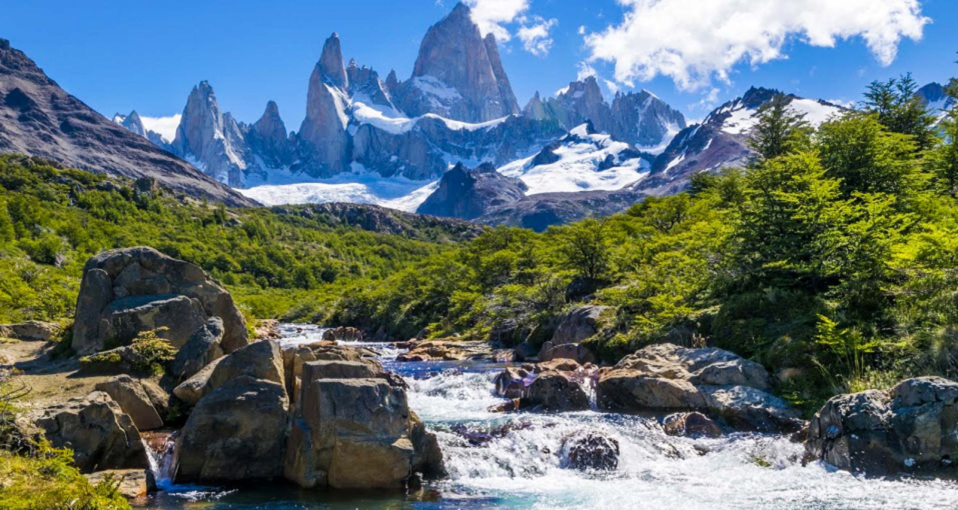 A clear stream flows over rocks in the foreground with green trees on both sides, leading to dramatic snow-capped mountains under a bright blue sky—a breathtaking scene along the Carretera Austral on your Patagonia Tour.