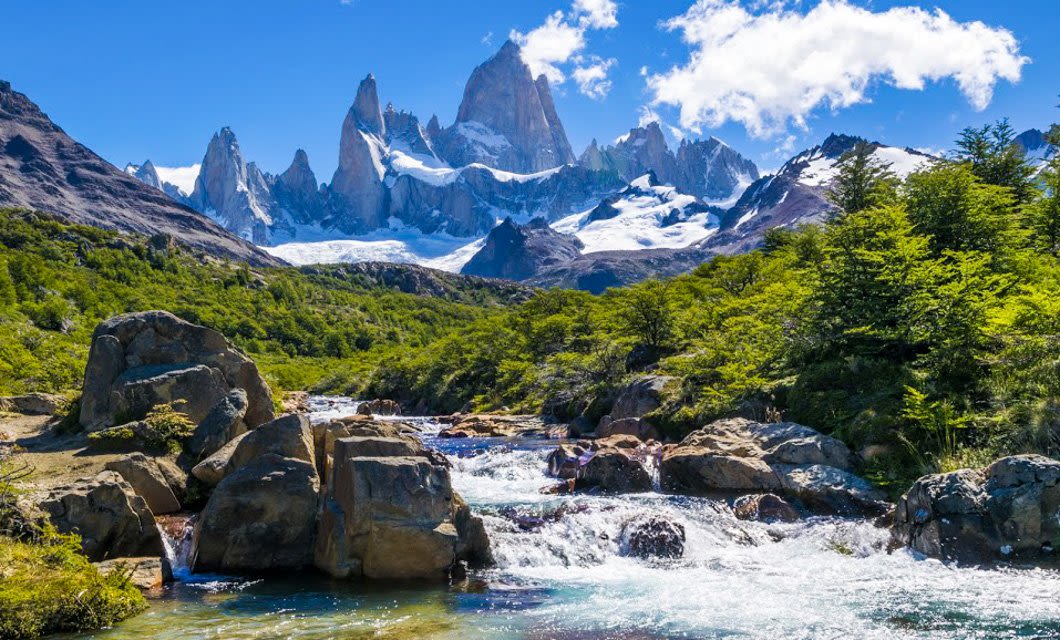 A clear mountain stream flows over rocks, surrounded by lush green trees. Snow-capped jagged peaks rise in the background under a bright blue sky—capturing the breathtaking beauty that defines the best of Argentina.