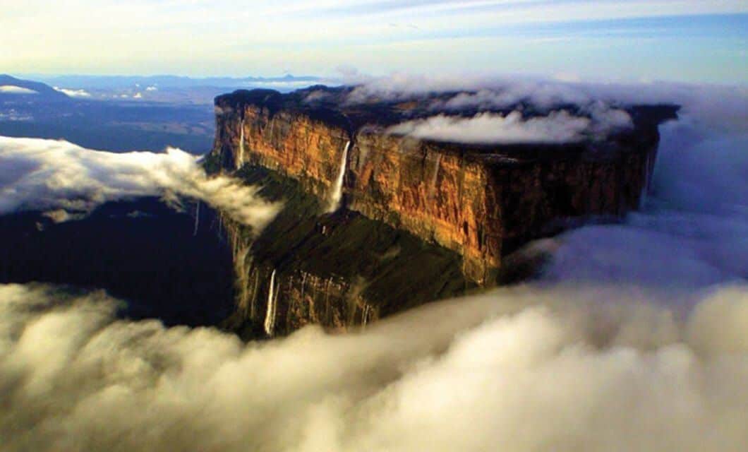 A dramatic, flat-topped mountain—Mount Roraima—rises above the clouds, with waterfalls cascading down its steep cliffs and mist surrounding the plateau. The landscape stretches out in the background, inviting adventurers to trek under a blue sky.