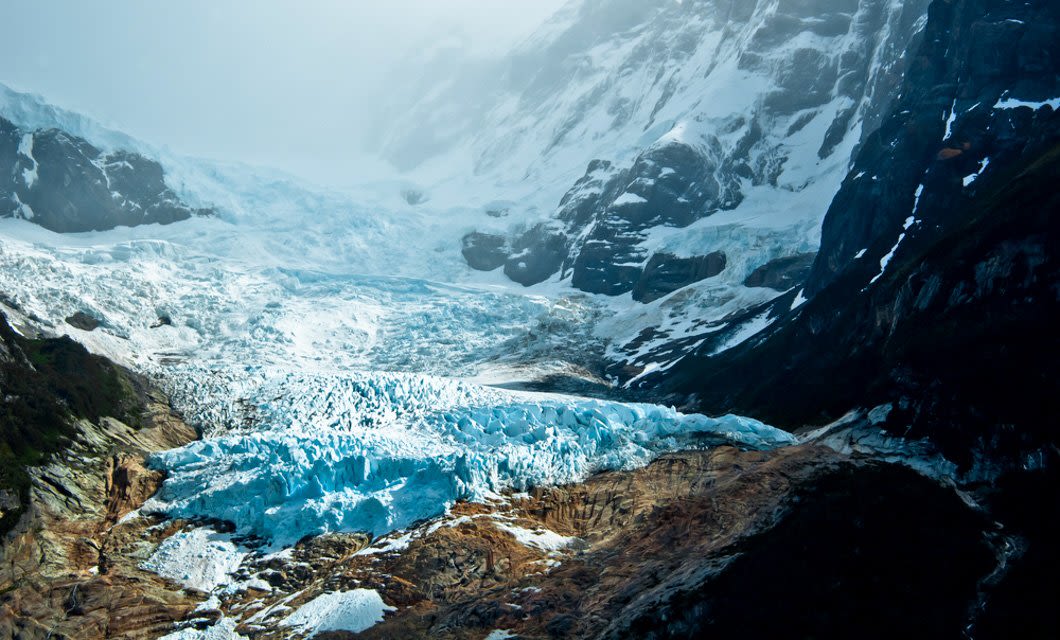 A glacier with blue-tinted ice stretches between rocky, snow-covered mountains under a cloudy Patagonian sky, with dark slopes and patches of greenery—an unforgettable scene on any Chile Tour or Glaciers Cruise.