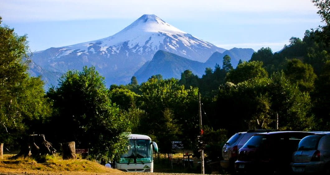 Snow-capped volcano rises above forested hills, with parked cars and a bus in the foreground under a clear blue sky—an iconic scene along the Lake Crossing route from Argentina to Chile.