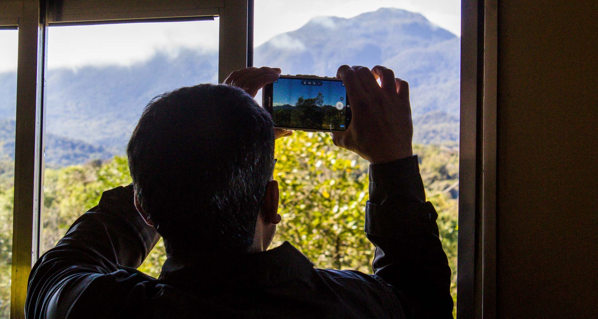 A person stands by a window, holding up a smartphone to capture the scenic mountain landscape along the Brazil Gold Route, with trees in the foreground and sunlight illuminating the stunning view.