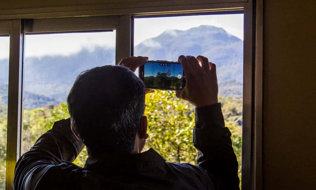 A person stands indoors, holding up a smartphone to take a photo of scenic mountain waterfalls visible through a window on a bright day.