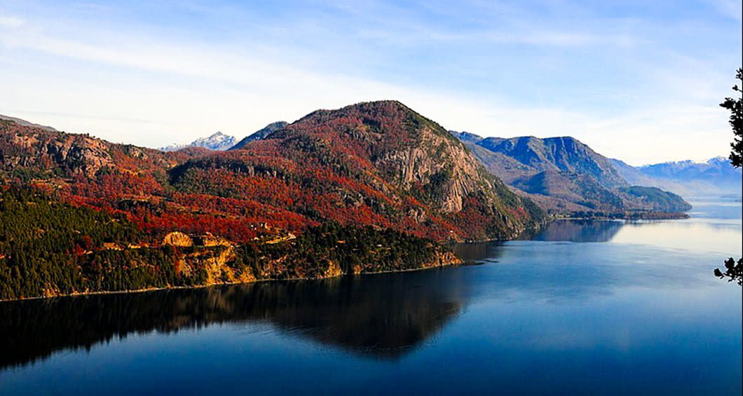 A calm blue lake, part of the Argentina to Chile Lake Crossing, is bordered by forested hills with autumn foliage in reds and greens, set against a backdrop of mountains under a partly cloudy sky.