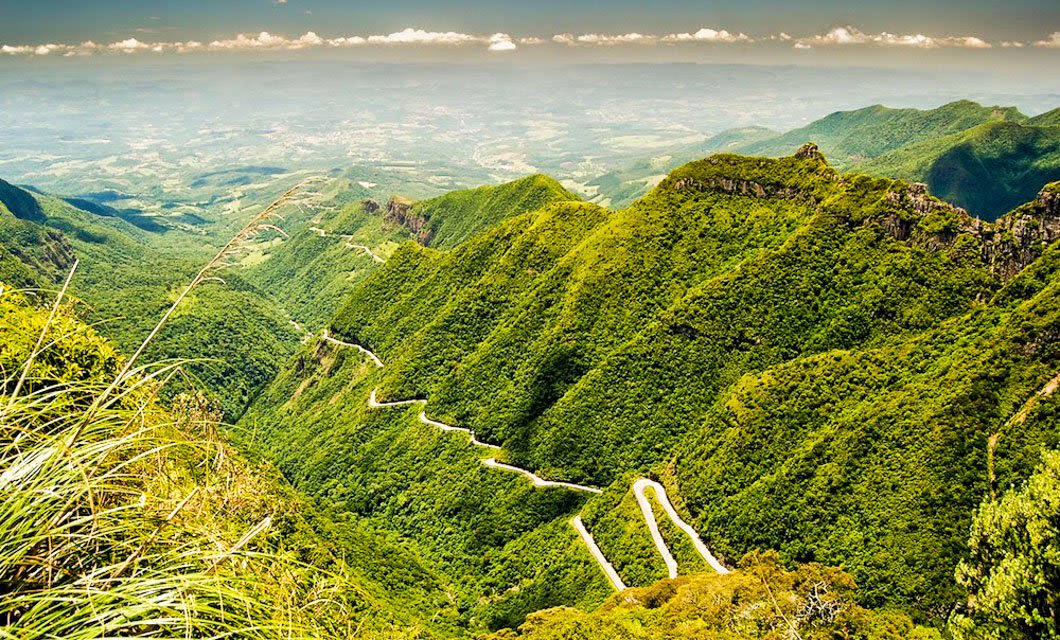 A winding road snakes through lush green mountains in Southern Brazil under a partly cloudy sky, with distant flatlands visible on the horizon—a perfect backdrop for adventure and horseback riding.