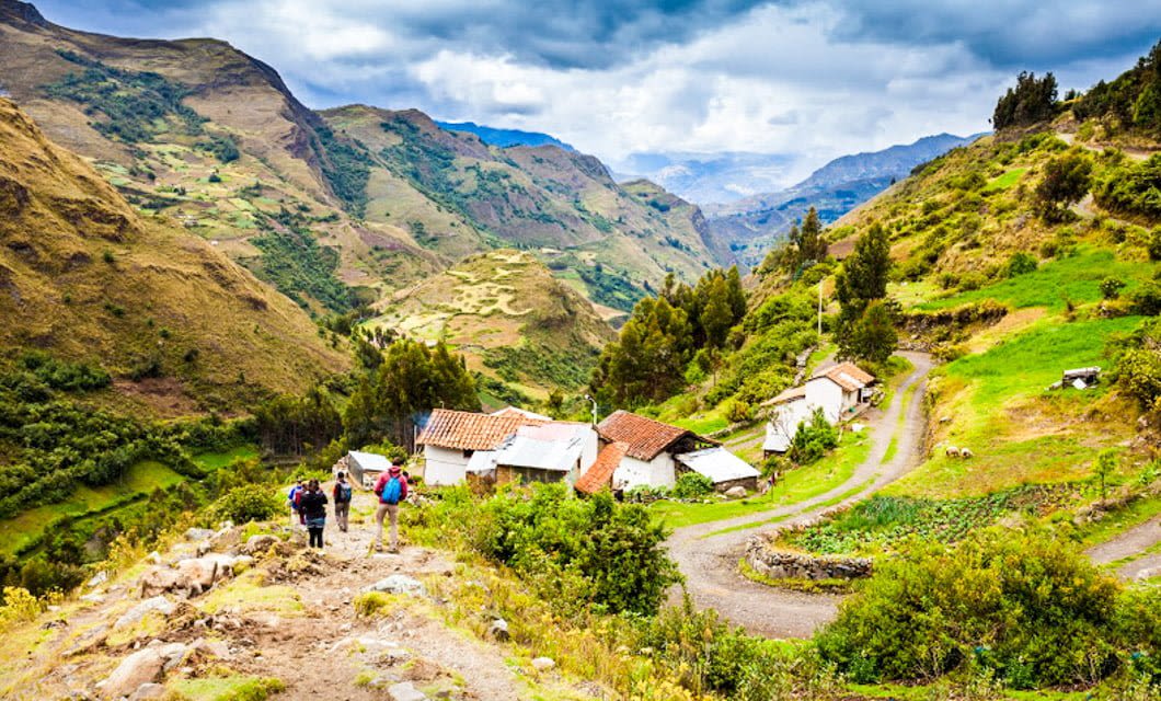 Hikers with backpacks walk down a lush, green mountain path past small houses in a scenic Huaraz valley, surrounded by the dramatic hills and winding roads of Peru Trek’s stunning Cordillera Blanca.