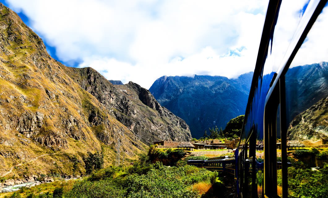 A scenic view of a train traveling through lush green valleys and rugged mountains under a partly cloudy sky, with the train’s reflection visible in the window—perfect for a Coast to Coast Tour adventure.