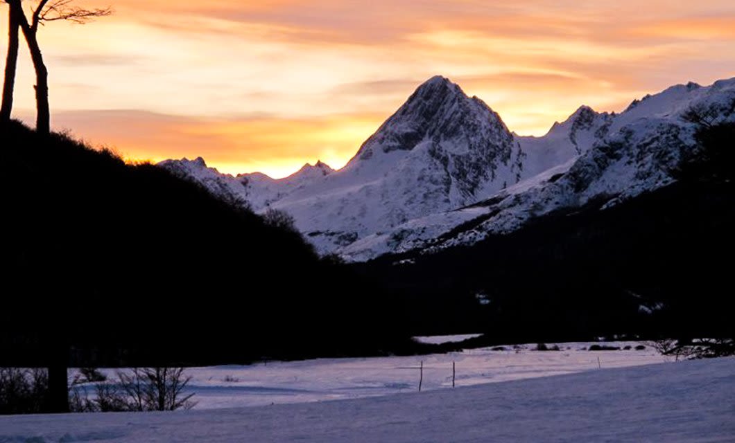 Snow-covered mountains under a dramatic orange and yellow sunset sky, with dark silhouettes of trees and hills in the foreground—an awe-inspiring scene perfect for those dreaming of skiing in Ushuaia across a stunning, partially frozen landscape.