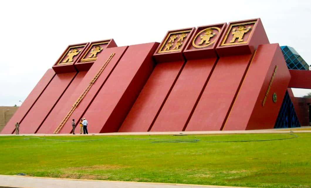 A large, modern red building with slanted walls and gold ancient motifs atop, set on green grass with people standing nearby—an iconic symbol of Chiclayo’s rich Cajamarca archaeology.