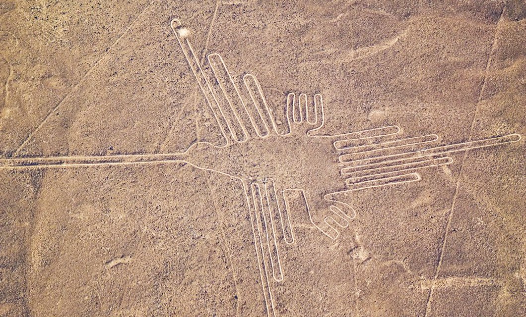 Aerial view of a large geoglyph in the shape of a hummingbird etched into the desert surface, known as one of the Nazca Lines in Peru—a must-see travel destination that highlights ancient Peruvian culture. The surrounding landscape is arid and flat.