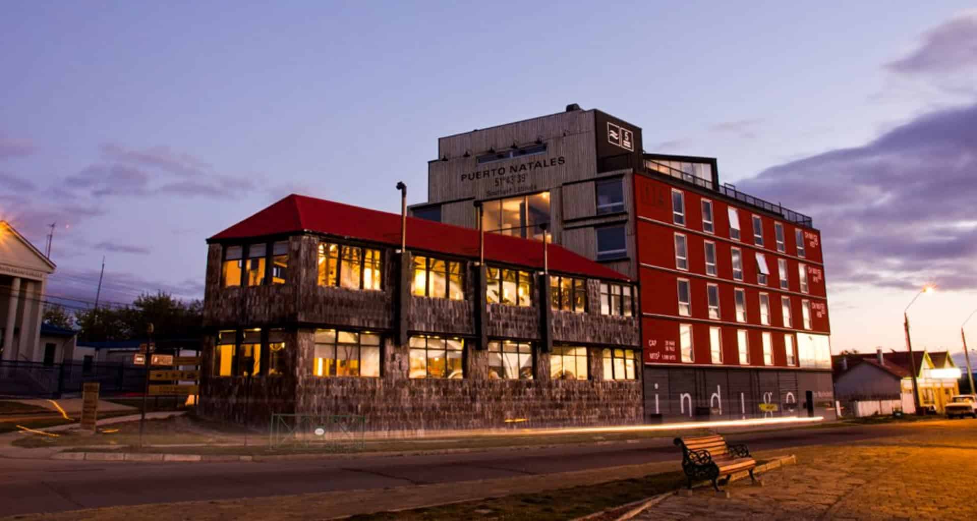 A modern building with a red roof and large windows glows with lights at dusk. The sign reads Puerto Natales, home to Noi Indigo Patagonia. The sky is purple, and a bench sits on the sidewalk in the foreground.