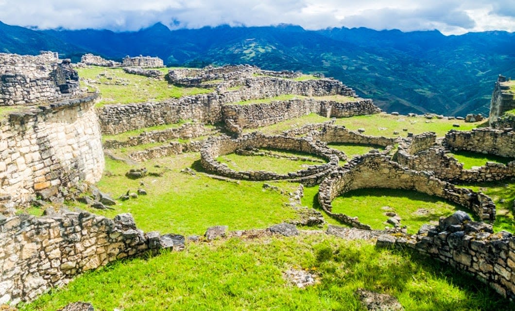 Ancient stone ruins with circular and rectangular foundations sit on a grassy hilltop near Chachapoyas, surrounded by green mountains under a cloudy sky.